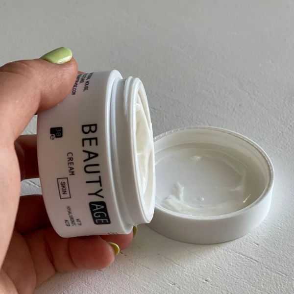 Mano que inclina el tarro blanco abierto de Beauty Age Skin, con la crema a la vista y la tapa a su lado