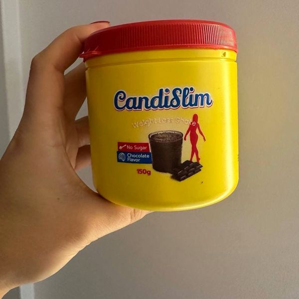 Mano que sostiene el bote amarillo de CandiSlim, de 150 g y tapa roja, con un batido de chocolate dibujado