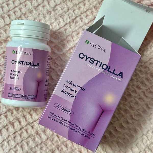 Bote blanco de Cystiolla tumbado junto a su caja rosa y lila abierta, sobre una tela de color crema