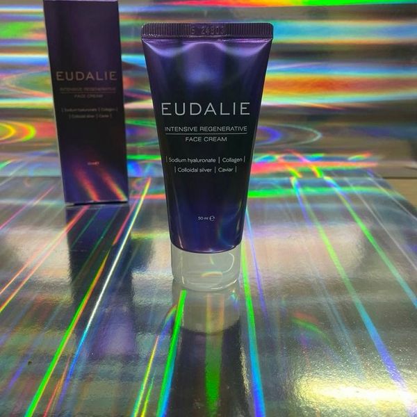 Tubo violeta de Eudalie, de 50 ml, apoyado en su tapón transparente junto a la caja, sobre un fondo irisado