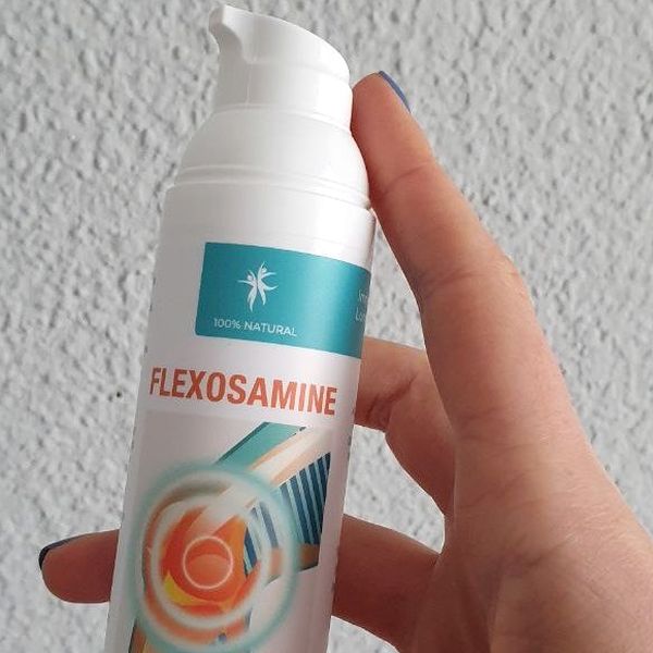 Mano que sostiene el frasco blanco con dosificador de Flexosamine, de franja turquesa, ante una pared gris
