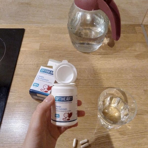 Mano que sostiene el bote de Optiheart abierto junto a un vaso de agua