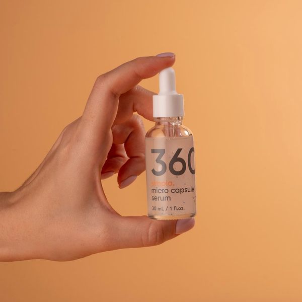 Mano que sostiene el frasco cuentagotas de Simpla 360, sérum de microcápsulas de 30 ml, sobre fondo naranja