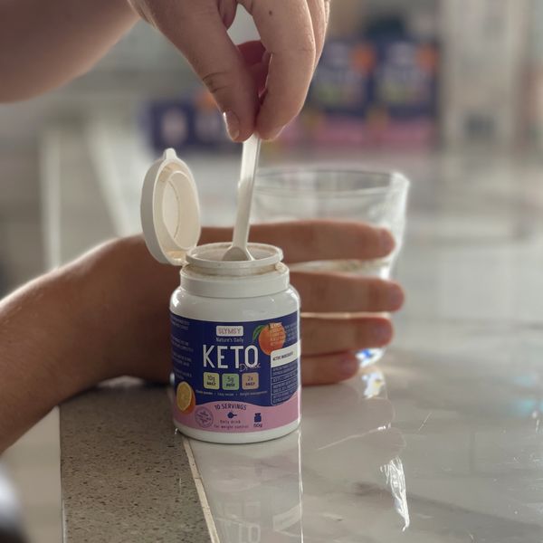 Cacito de Slymsy Keto Drink sobre el bote, con un vaso de agua detrás