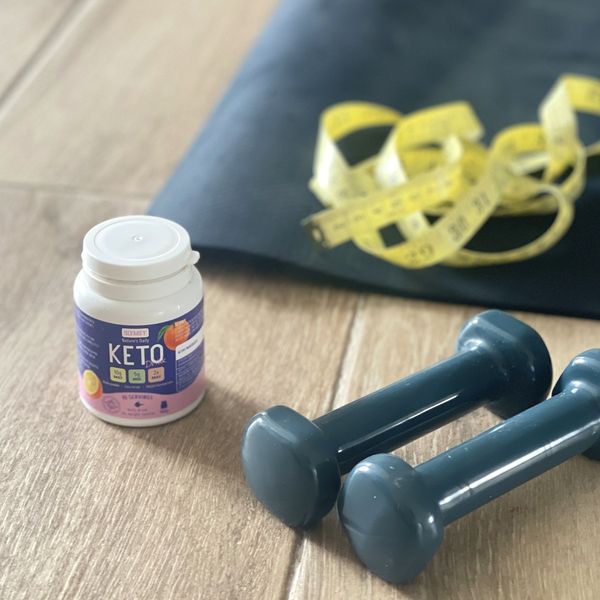 Bote de Slymsy Keto Drink en el suelo junto a unas mancuernas y una cinta métrica